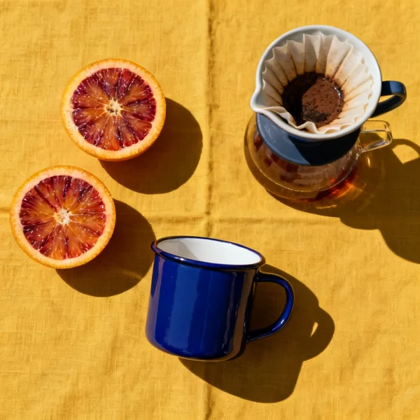 Top-down flat lay of a ceramic pour-over coffee set on a mustard yellow linen cloth, a cobalt blue enamel mug, two halved blood oranges, hard noon sunlight with crisp shadows, saturated color blocking, square social tile composition